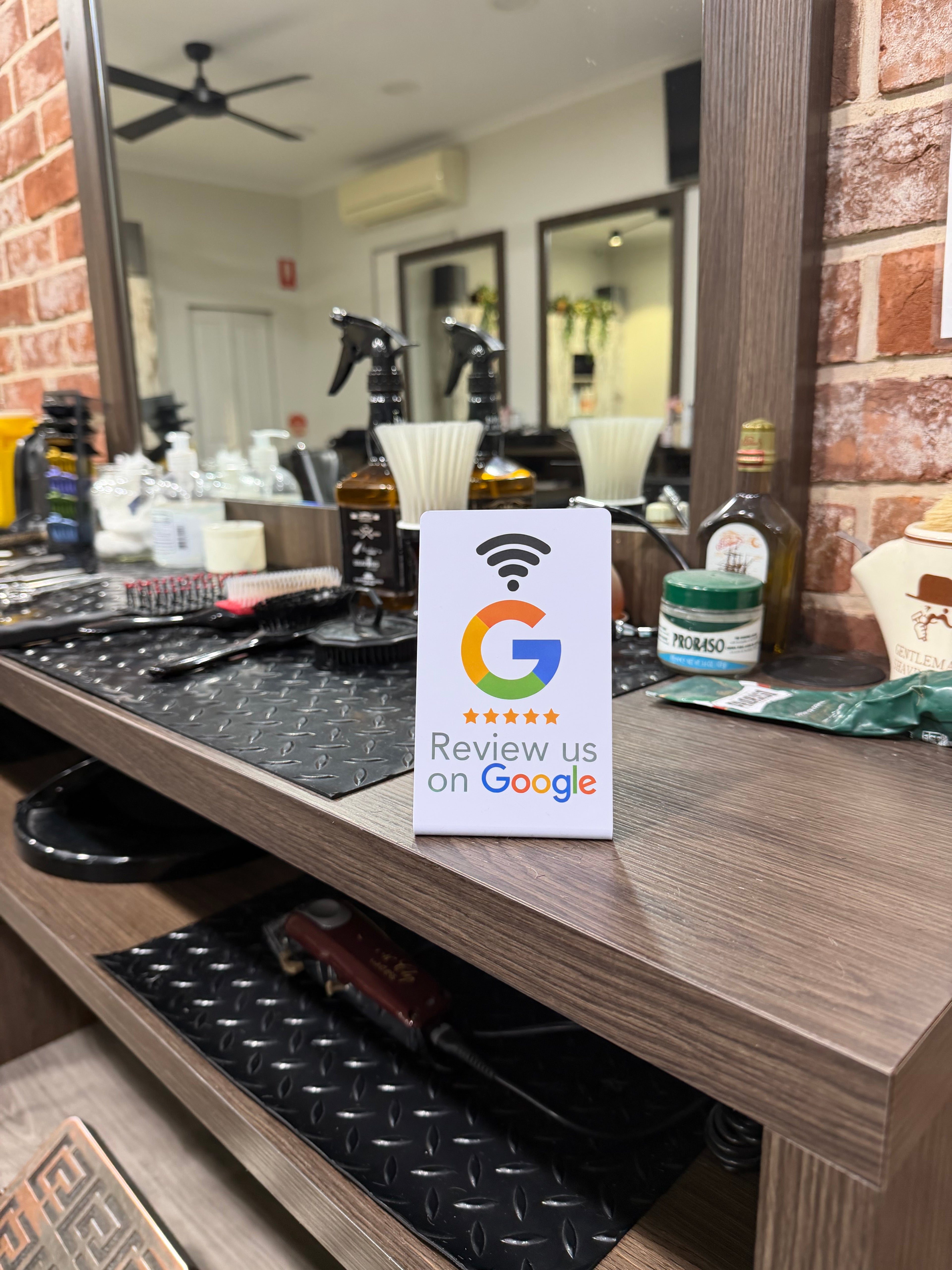 A white Google review card standing on a wooden counter in a salon or beauty shop, with spray bottles, hair products, mirrors, and a brick-pattern wall in the background.
